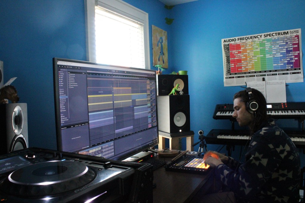 Photo of a man sitting at a desk with a large monitor. He is wearing headphones and making music on a beat machine.