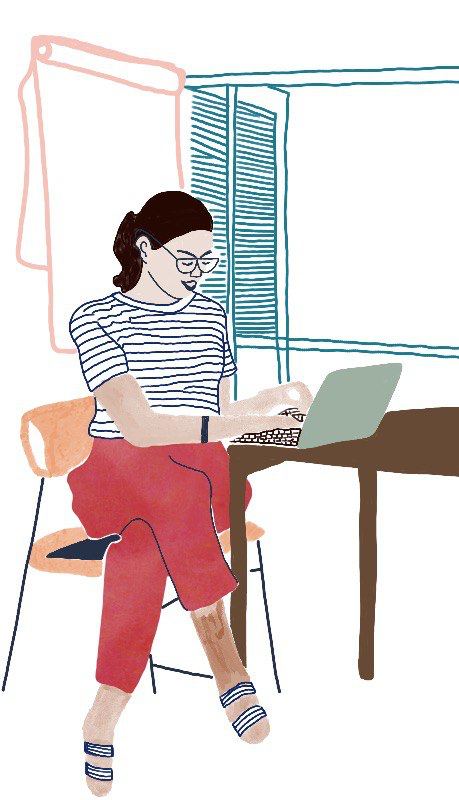 Eine Frau sitzt vor einem geöffneten Fenster mit übergeschlagenen Beinen auf einem Stuhl an einem Tisch und tippt in einen Laptop