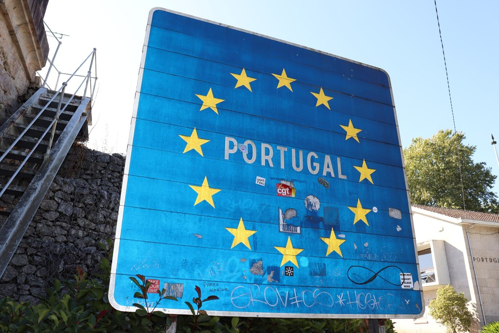 A photograph of a Portugal border sign, which features the country's name on the blue field and surrounded by the yellow stars of the European Union flag, and has some weathered stickers and graffiti on it