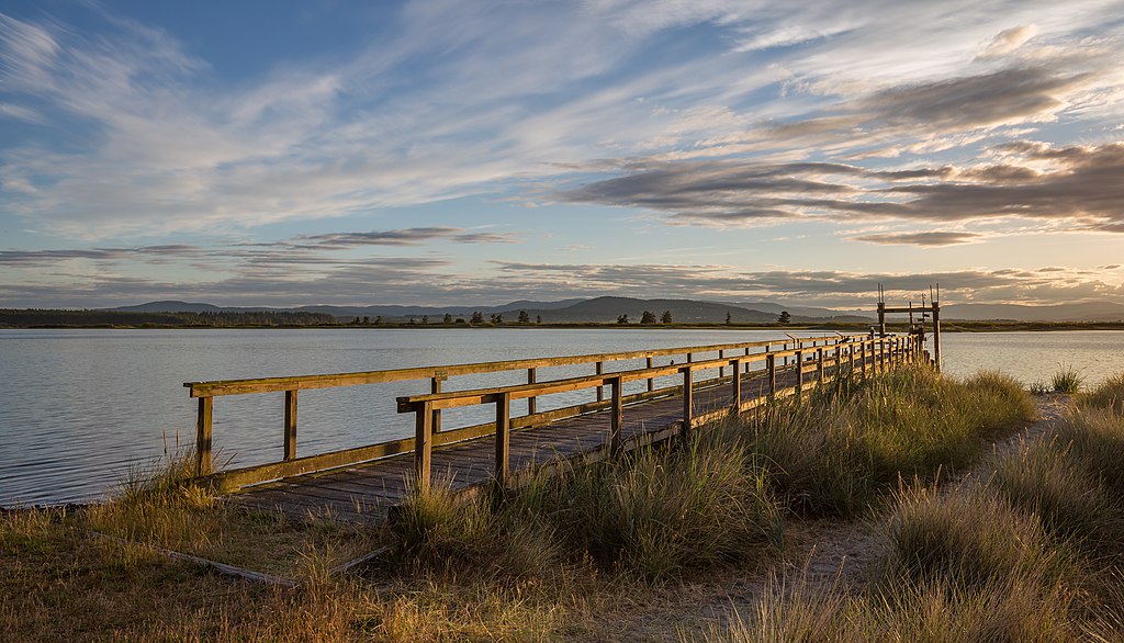 A small wooden pier surrounded by clumps of grass on the right and water on the left is lit up by the light of the setting sun. The sky is full of wispy clouds with hills and trees visible in the distance on the water's edge.