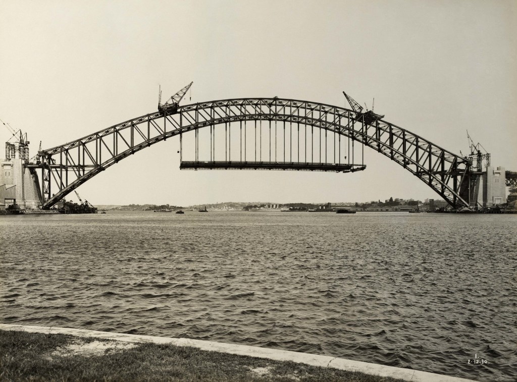 A black and white photograph of the Sydney Harbour Bridge under construction, as viewed from McMahon's Point