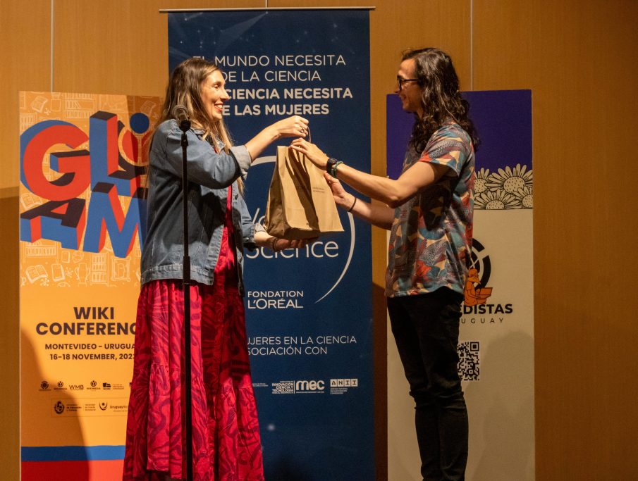 A woman stands on the stage with a package in her hands and delivers it to a man