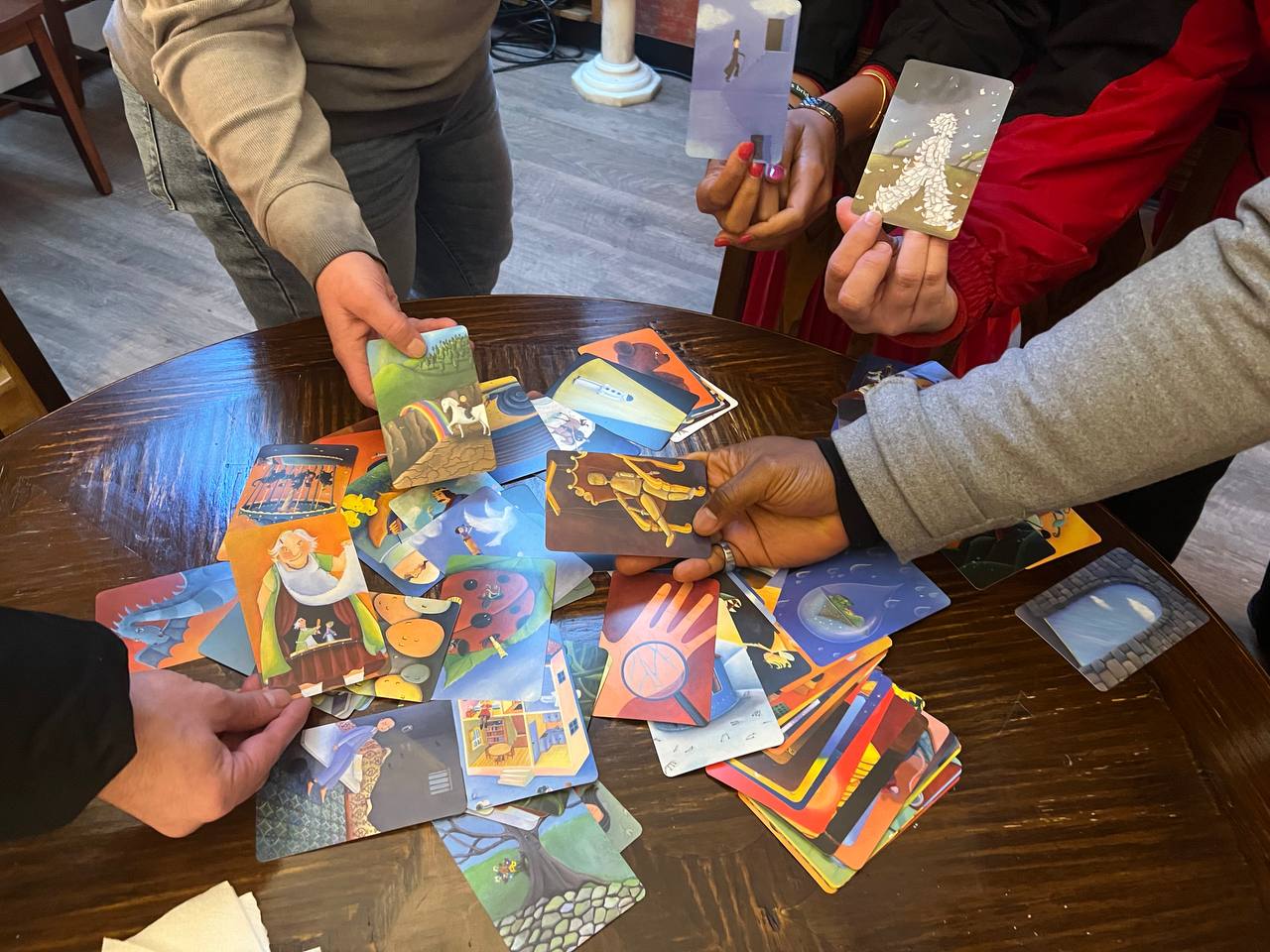 5 different hands, all holding a conversation card above a pile of unpicked conversation cards. The conversation cards have different drawings on them.