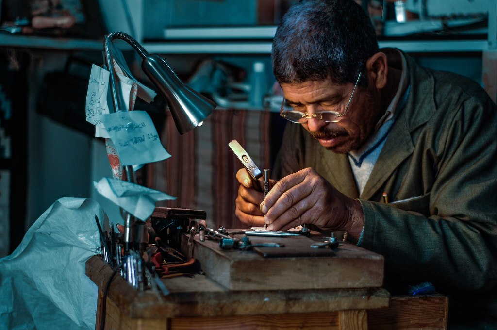 An engraver hunches over detailed work in his busy workshop. A small light highlights the intricate work he is doing.